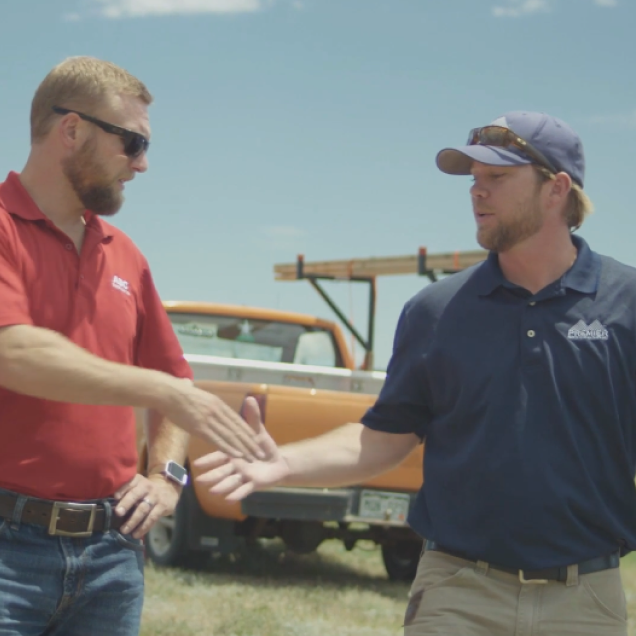 Two white men shaking hands in front of a pickup truck on a job site. One wears a red polo and sunglasses, and the other is in a navy polo and cap.